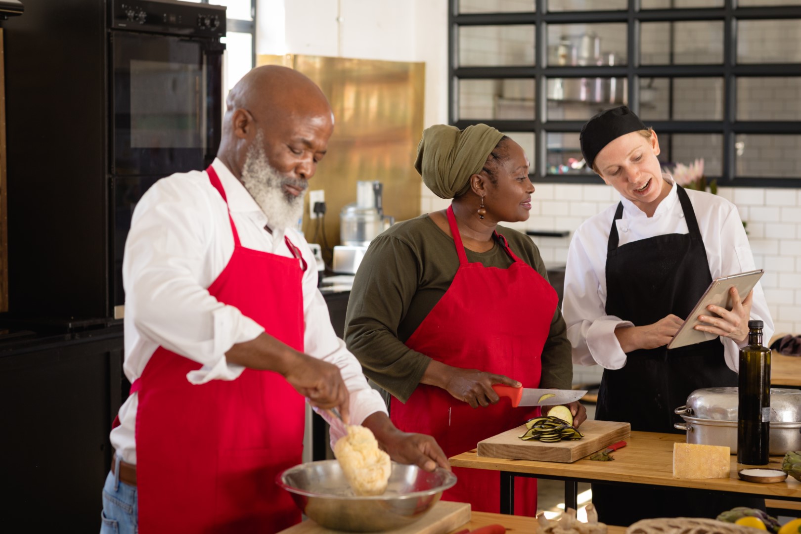 Side view of an Senior African American woman and man at a cookery class, listening to instructions from a Caucasian female chef wearing chefs whites and a black hat and apron, using a tablet computer while they work standing at a wooden table of ingredients